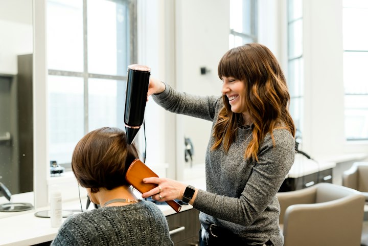 A stylist blow-dries a client's short hair while they both smile